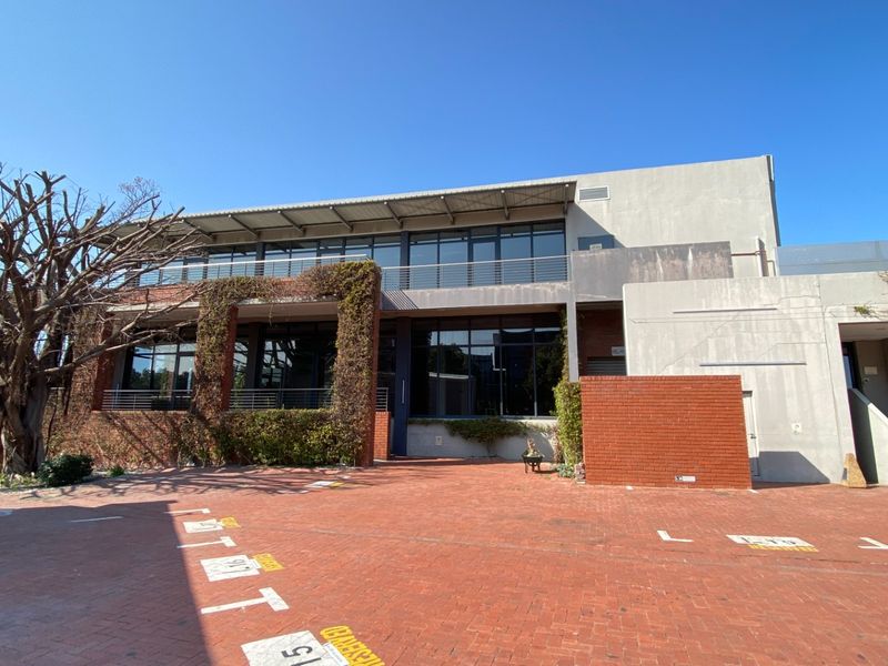 Front exterior view of a modern office building featuring large glass windows, a balcony, and brick paved parking area.