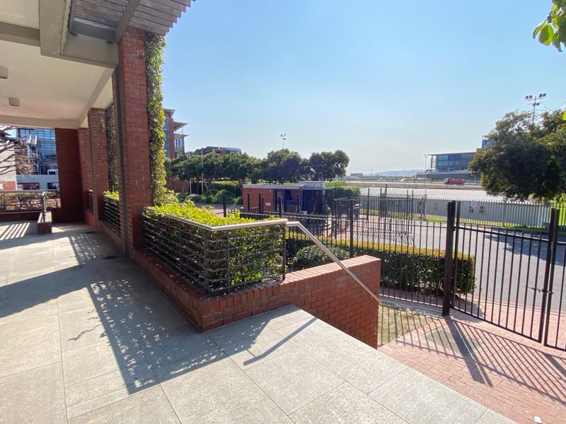 A covered outdoor patio area featuring brick pillars, tiled flooring, and metal security fencing overlooking the office park grounds.
