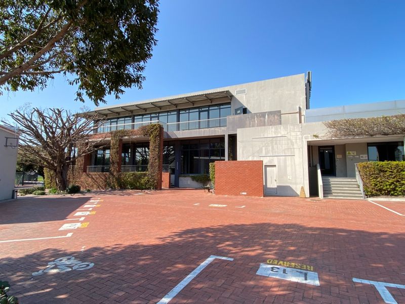Front view of a modern office building featuring a paved parking area, brick and concrete facade, and large glass windows with balcony access.