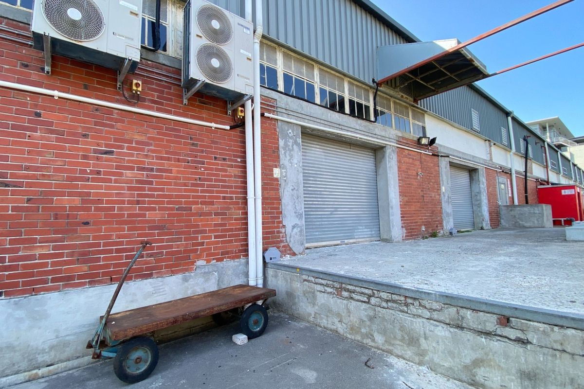 Rear exterior view of an industrial office building featuring a concrete loading dock, multiple roller shutter doors, and external air conditioning units.