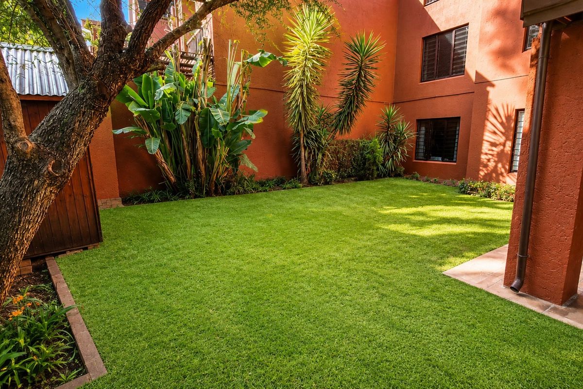 A well-maintained rear garden featuring a lush green lawn, paved patio area, and a timber storage shed against a terracotta-colored building facade.