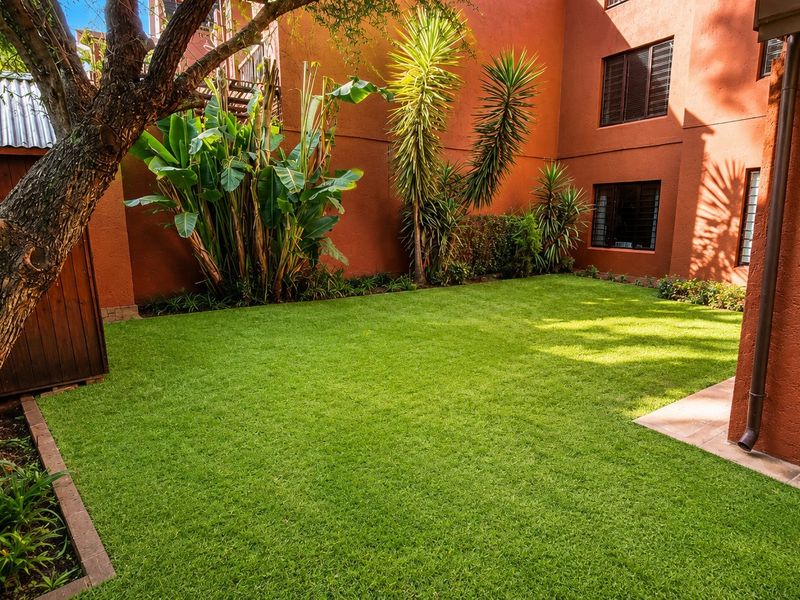 A well-maintained rear garden featuring a lush green lawn, paved patio area, and a timber storage shed against a terracotta-colored building facade.