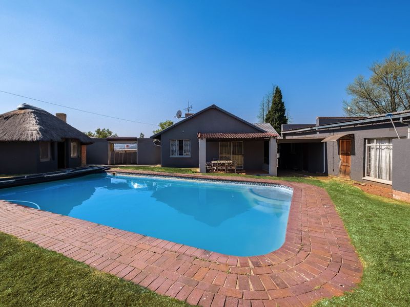 A backyard pool area featuring a swimming pool with brick coping, a covered patio, and a separate thatched roof structure.