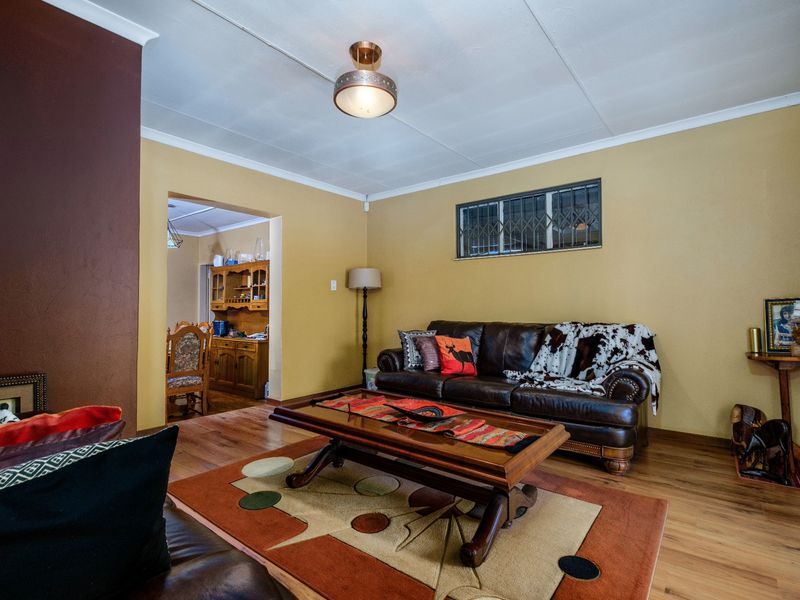 A comfortable living room featuring laminate flooring, a ceiling light fixture, and a window fitted with security bars, leading into an adjacent dining area.