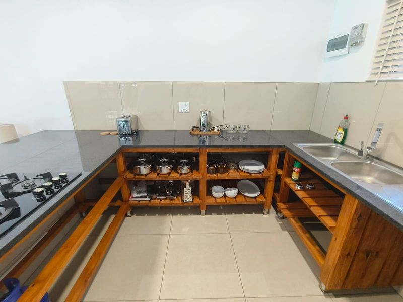 A functional kitchen featuring granite countertops, a built-in gas hob, double stainless steel sink, and open timber shelving for storage.