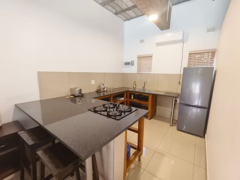 A functional kitchen featuring a dark stone countertop, integrated gas hob, stainless steel sink, and tiled splashback, complemented by modern tiled flooring.