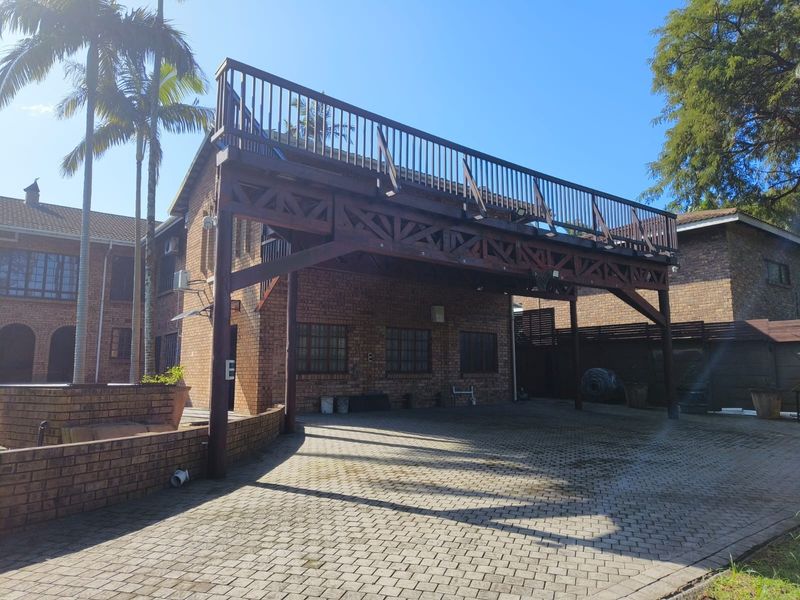 Rear exterior view of the cottage featuring a large elevated timber deck supported by exposed beams, overlooking a spacious brick-paved driveway area.