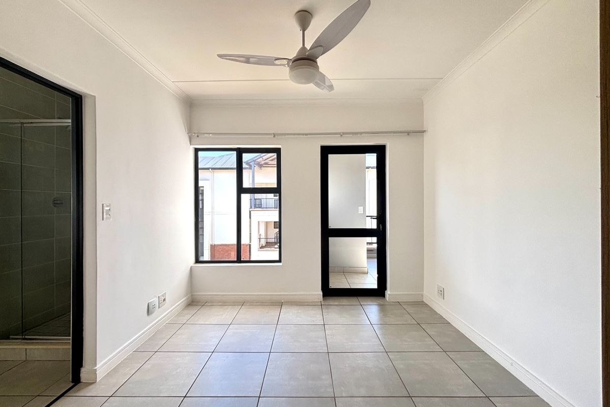 A bright, empty bedroom featuring neutral tiled flooring, a ceiling fan, and direct access to an outdoor balcony through an aluminium-framed door.