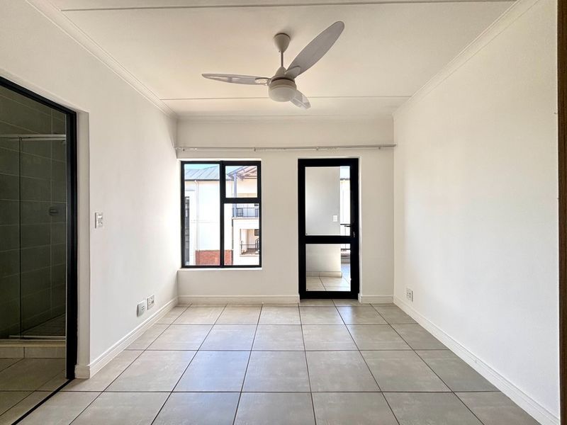 A bright, empty bedroom featuring neutral tiled flooring, a ceiling fan, and direct access to an outdoor balcony through an aluminium-framed door.