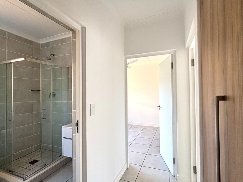 A bright hallway featuring tiled flooring, providing access to a modern bathroom with a glass shower enclosure and a bedroom beyond.