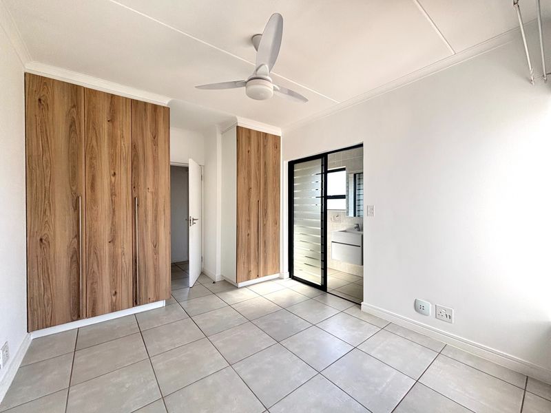 A bright, modern bedroom featuring neutral tiled flooring, ample built-in wooden cabinetry, a ceiling fan, and direct access to an en-suite bathroom.