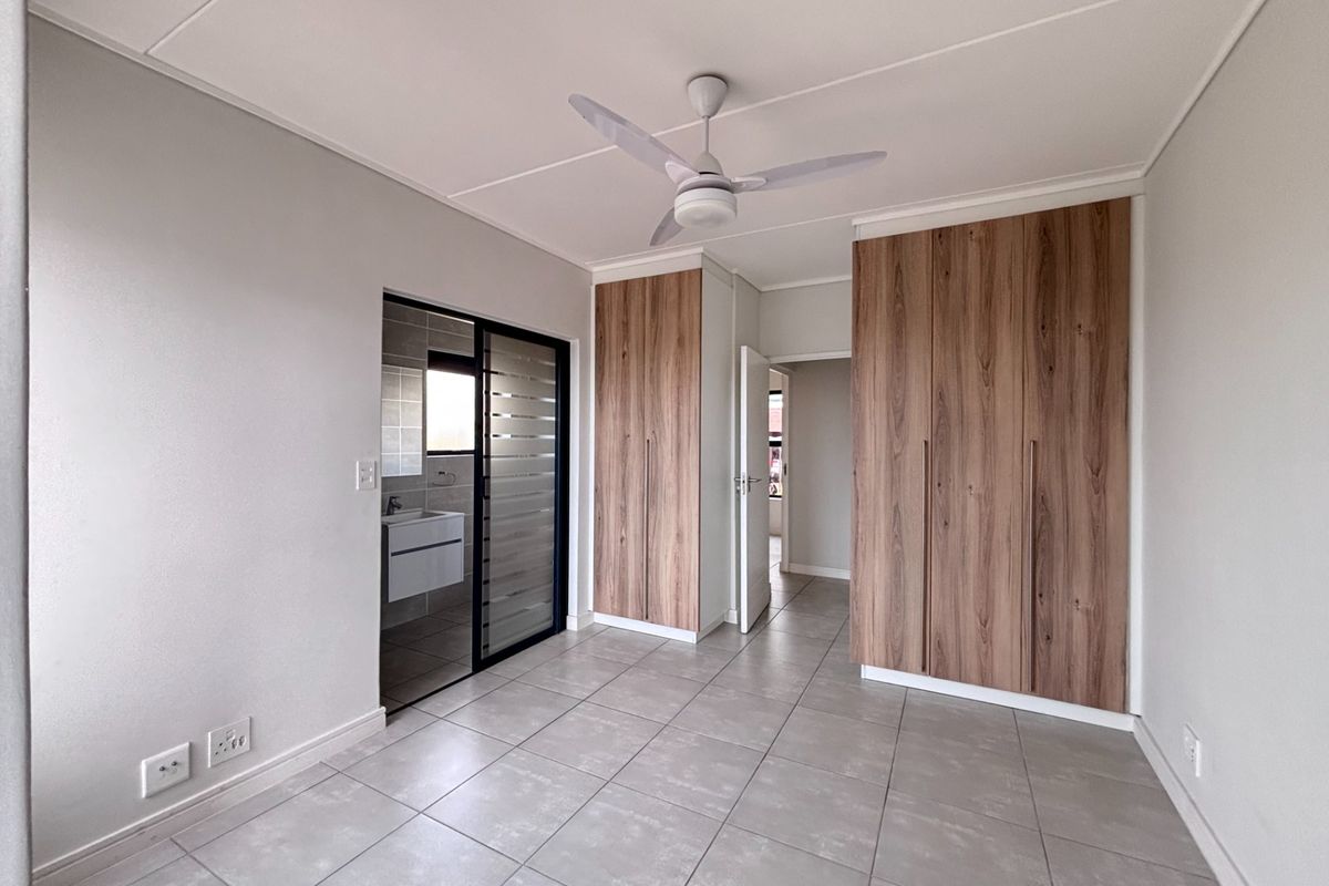 A bright, modern bedroom featuring neutral tiled flooring, built-in wood-finish cupboards, a ceiling fan, and direct access to an en-suite bathroom.