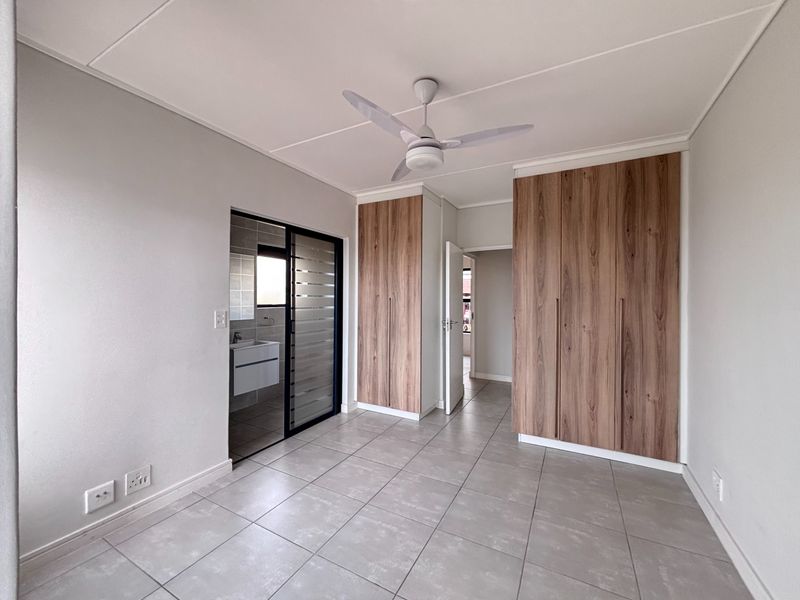 A bright, modern bedroom featuring neutral tiled flooring, built-in wood-finish cupboards, a ceiling fan, and direct access to an en-suite bathroom.