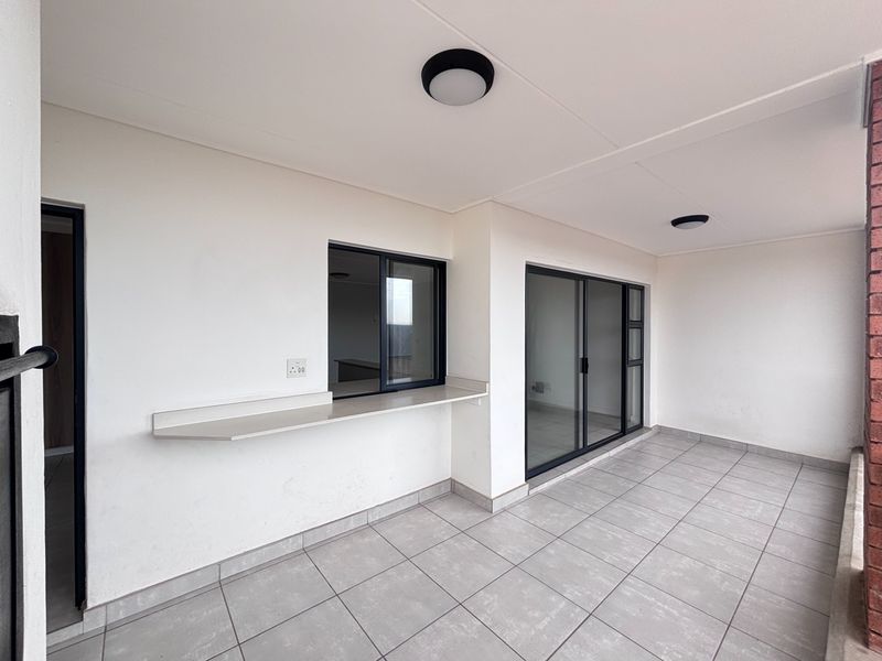 A clean, covered balcony featuring tiled flooring, a convenient serving counter, and sliding glass doors providing access to the interior living space.