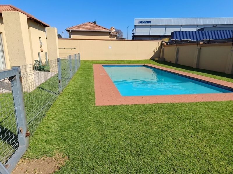 A well-maintained outdoor pool area featuring a rectangular swimming pool with brick coping, surrounded by a lawn and secure perimeter fencing.