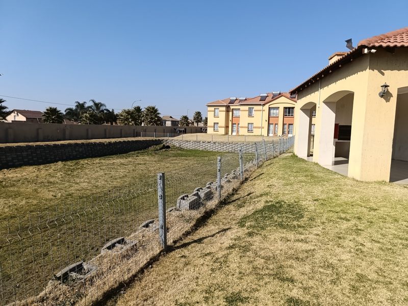 Rear view of the apartment complex featuring a covered patio with a built-in braai, overlooking a fenced garden area with a retaining wall.