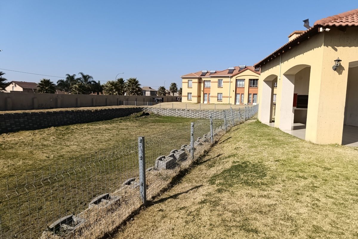 Rear view of the apartment complex featuring a covered patio with a built-in braai, overlooking a fenced garden area with a retaining wall.