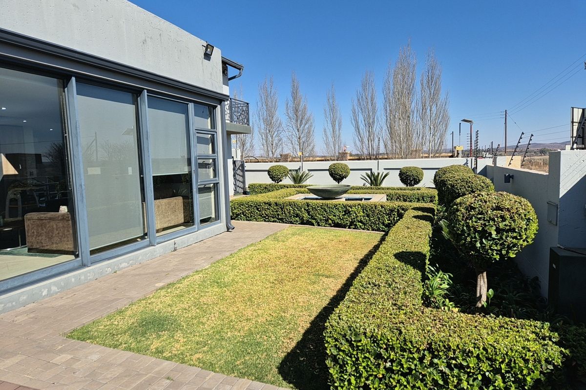 A well-maintained rear garden featuring a paved patio, manicured hedges, a central water feature, and large sliding glass doors leading into the home.