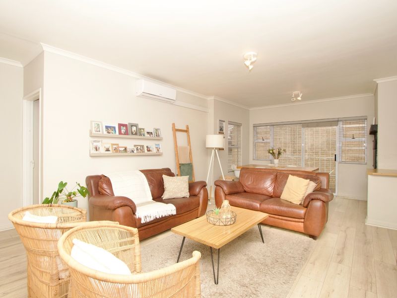 A well-maintained living room featuring light-toned laminate flooring, a wall-mounted air conditioning unit, and large windows providing natural light.