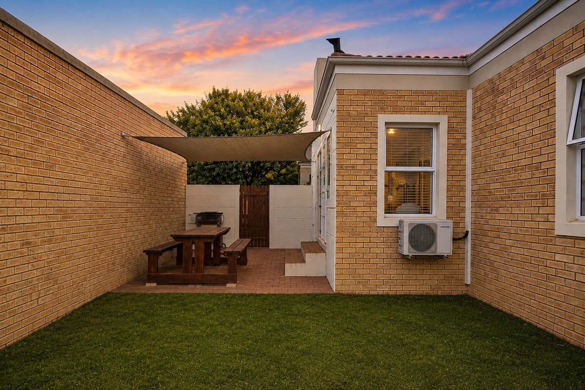 A well-maintained rear courtyard featuring a paved patio area with a shade sail, brick exterior walls, and a small private lawn.