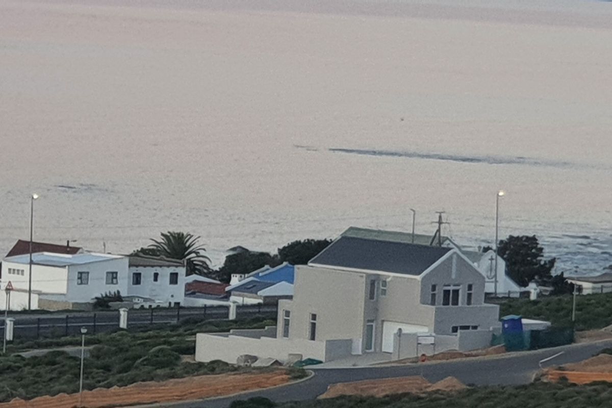 An elevated aerial view of a coastal residential property featuring a modern home with a pitched roof, driveway, and surrounding neighborhood landscape.
