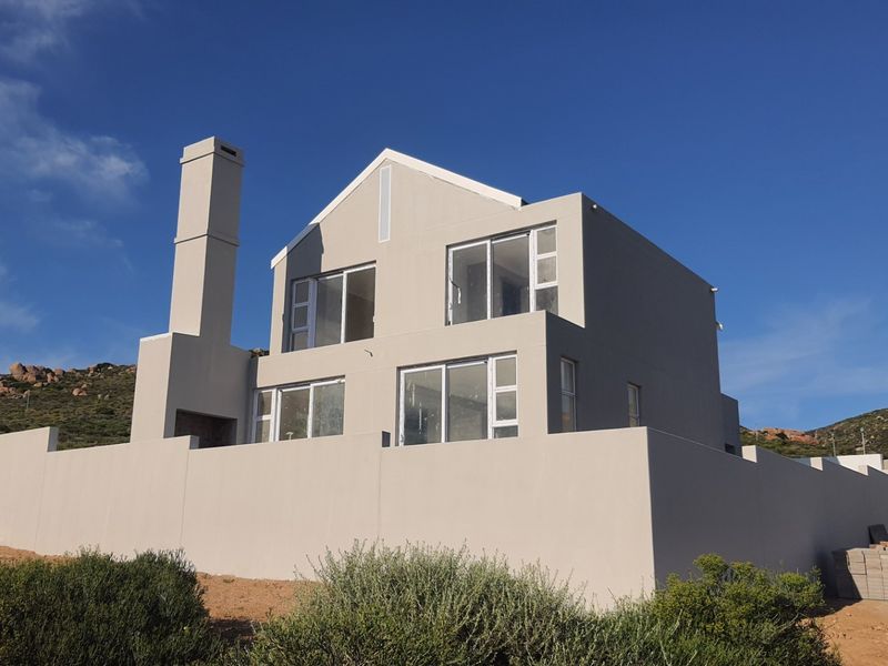 A modern two-story house exterior featuring a prominent chimney, large sliding glass doors, and a surrounding perimeter wall in a coastal setting.