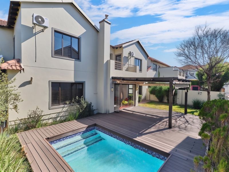 Rear exterior view featuring a swimming pool, timber decking, and a pergola, complemented by a two-story residential facade with an upper-level balcony.