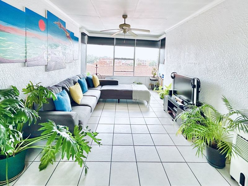 A bright living room featuring tiled flooring, a ceiling fan, and large windows providing natural light and an exterior view.