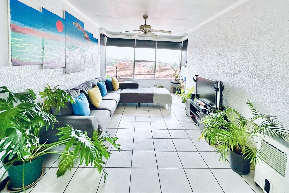 A bright living room featuring tiled flooring, a ceiling fan, and large windows providing natural light and an exterior view.