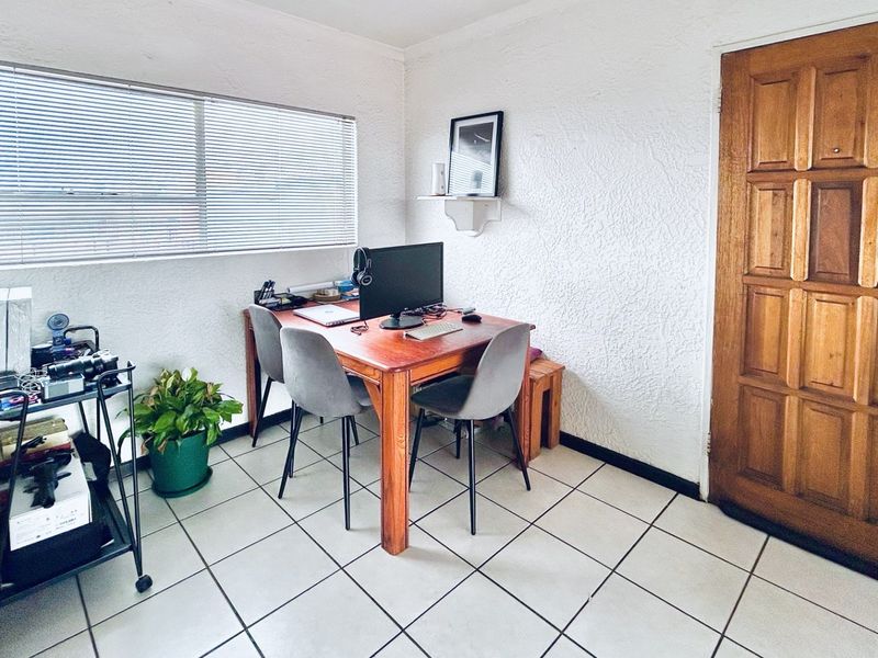 A functional home office space featuring tiled flooring, a large window with venetian blinds, and a solid timber panel door.