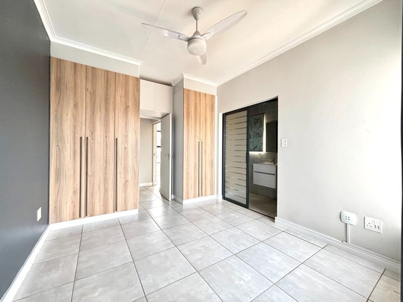 A bright, modern bedroom featuring neutral tiled flooring, ample built-in wooden cupboards, a ceiling fan, and direct access to an en-suite bathroom.