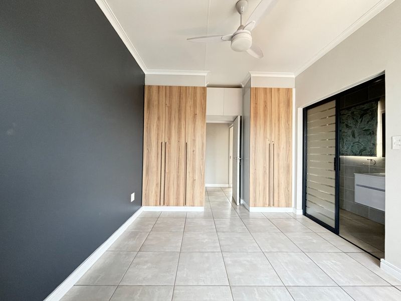 A bright, modern bedroom featuring neutral tiled floors, built-in wood-finish cupboards, a ceiling fan, and an en-suite bathroom access via a sliding door.