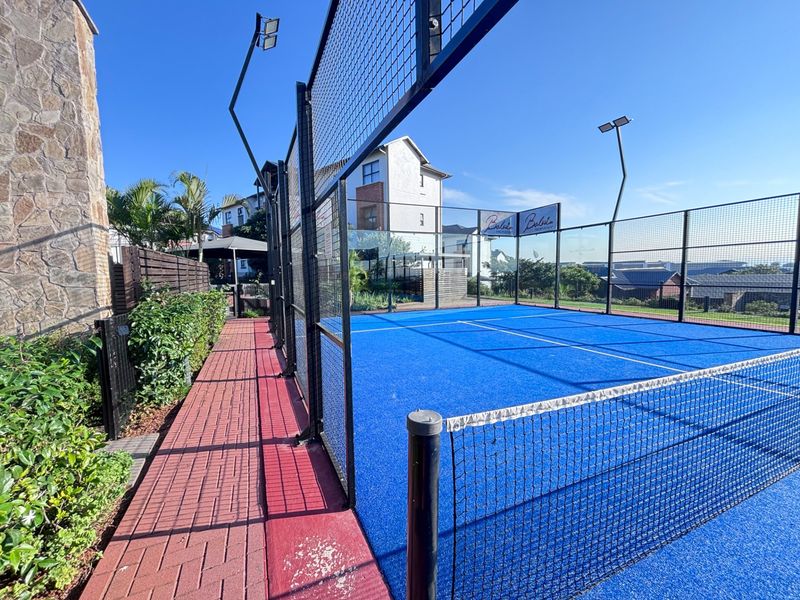 Well-maintained outdoor padel court featuring blue synthetic turf, perimeter fencing, and integrated floodlighting within the apartment complex.