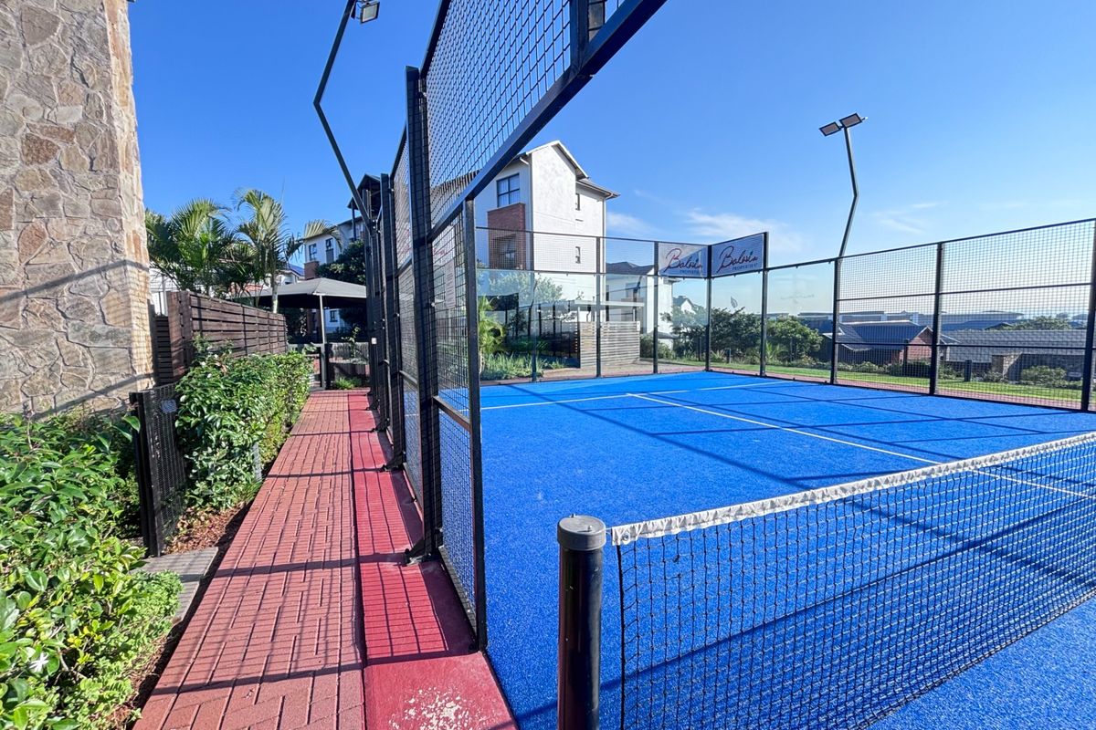 Well-maintained outdoor padel court featuring blue synthetic turf, perimeter fencing, and integrated floodlighting within the apartment complex.