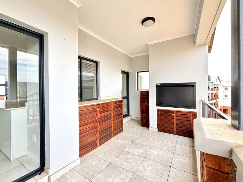 A spacious covered balcony featuring tiled flooring, built-in wooden cabinetry, and a built-in braai, offering a functional outdoor entertainment area.