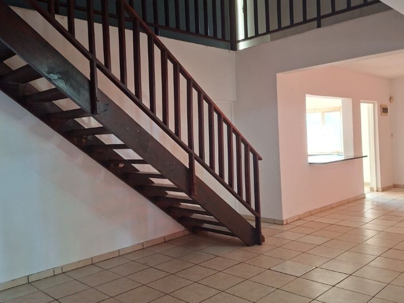 Spacious open-plan living area featuring a prominent wooden staircase, tiled flooring, and a convenient pass-through window connecting to the kitchen area.