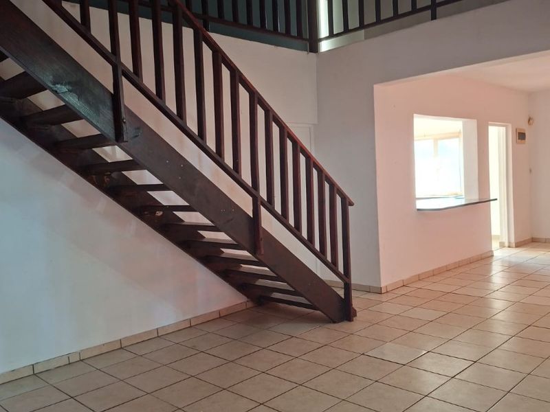 Spacious open-plan living area featuring a prominent wooden staircase, ceramic tiled flooring, and a convenient pass-through window to the kitchen area.
