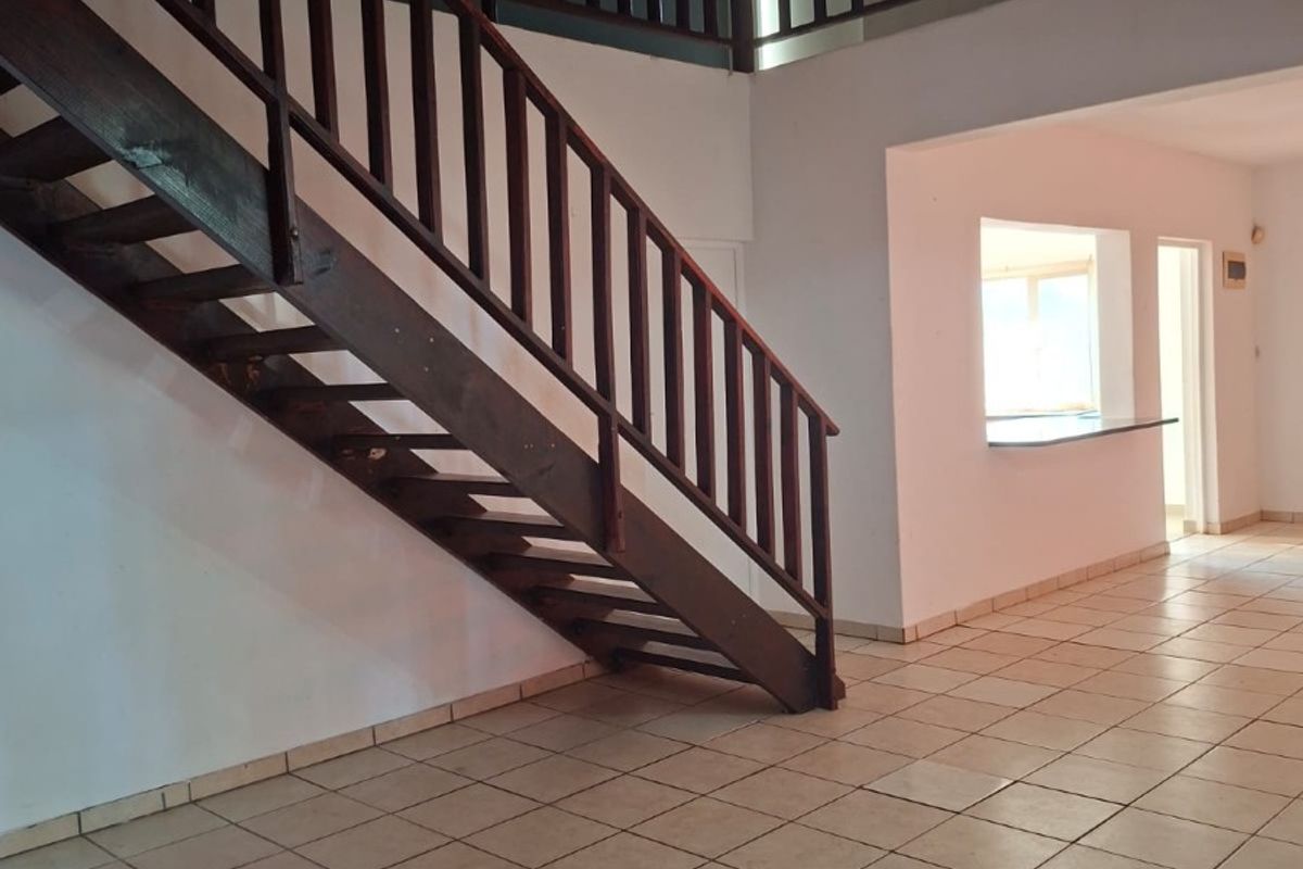 Spacious open-plan living area featuring a prominent wooden staircase, ceramic tiled flooring, and a convenient pass-through window to the kitchen area.