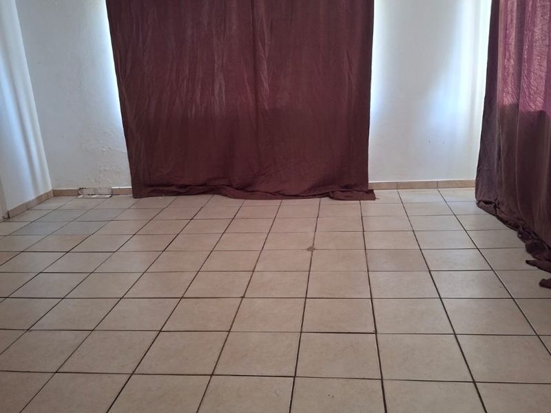 An empty living room featuring ceramic floor tiles and curtain rods, showing signs of wear on the flooring and wall surfaces.