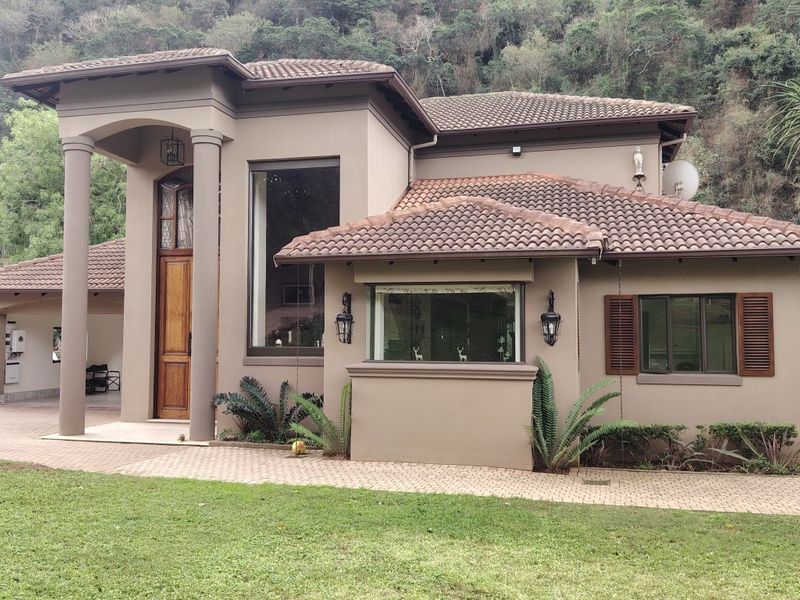 A well-maintained double-story house featuring a tiled roof, covered entrance with columns, and a paved driveway set against a natural hillside backdrop.