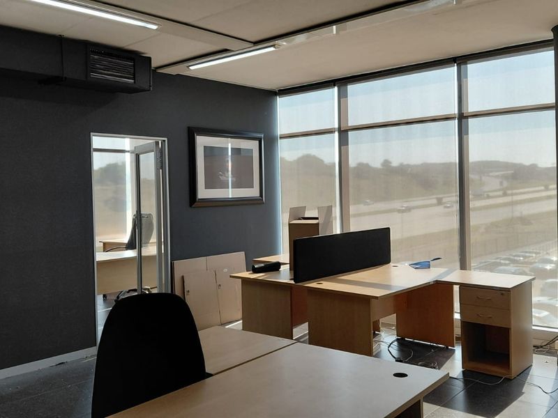 A modern office space featuring large floor-to-ceiling windows with natural light, dark accent walls, tiled flooring, and integrated ceiling lighting.