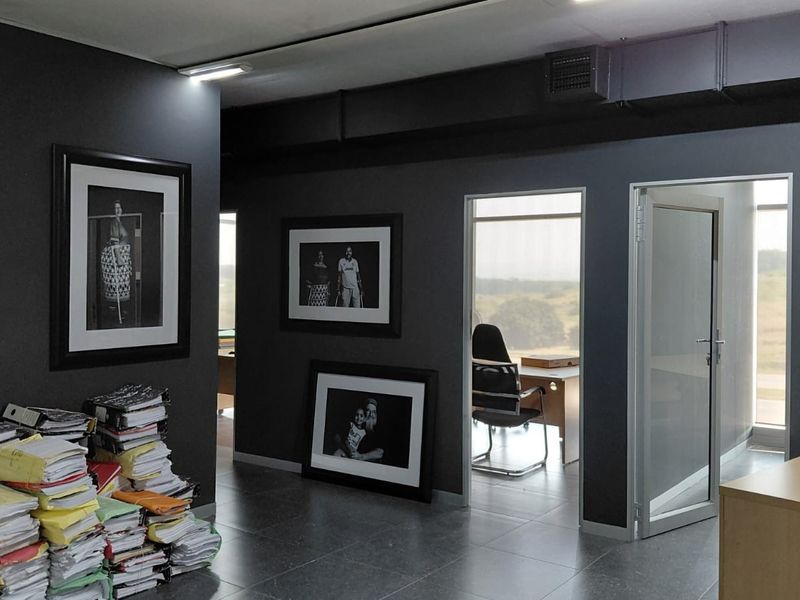 Modern office space featuring dark grey walls, tiled flooring, exposed ceiling ducting, and glass-panelled doors leading to adjacent workspaces.