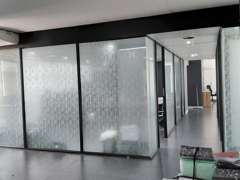 Modern office space featuring glass partition walls with privacy film, black metal framing, and tiled flooring throughout the corridor and partitioned work areas.