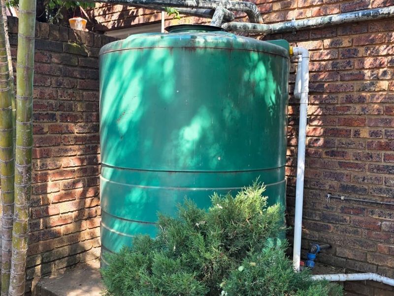 An outdoor utility area featuring a large green water storage tank installed against a brick boundary wall with associated plumbing infrastructure.