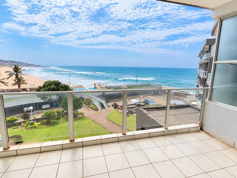 A spacious balcony featuring a modern glass balustrade and tiled flooring, offering expansive, unobstructed views of the ocean and coastline.
