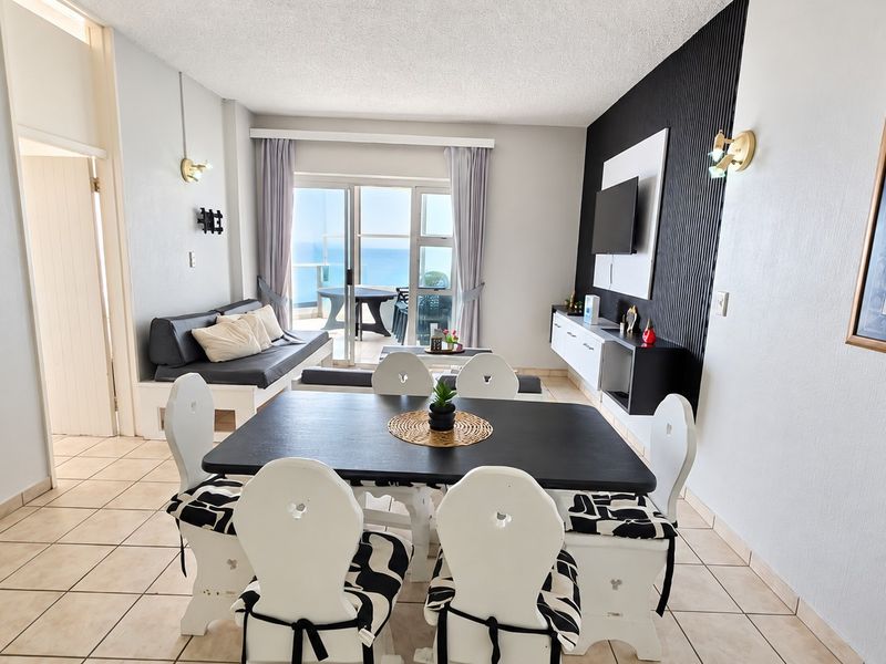 Open-plan living and dining area featuring tiled floors, a textured feature wall, and sliding glass doors leading to a balcony with ocean views.