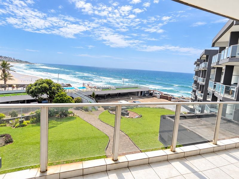 A spacious balcony featuring a modern glass balustrade and tiled flooring, offering expansive views of the ocean and surrounding coastal landscape.