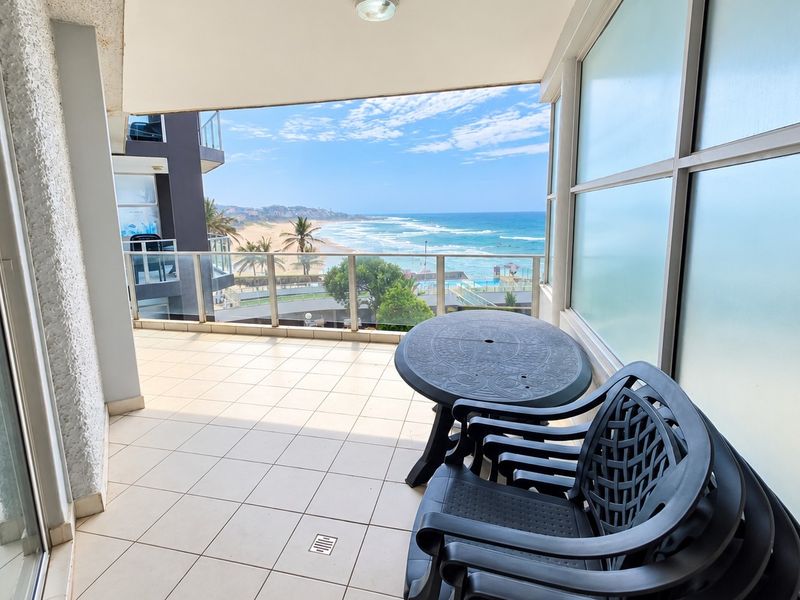 Covered balcony featuring tiled flooring, a glass balustrade, and a frosted glass privacy screen, offering expansive ocean and beach views.