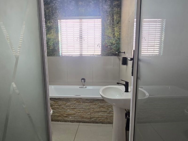 A functional bathroom featuring a pedestal basin, a built-in bathtub with stone-cladding detail, and tiled flooring, viewed through a frosted glass door.