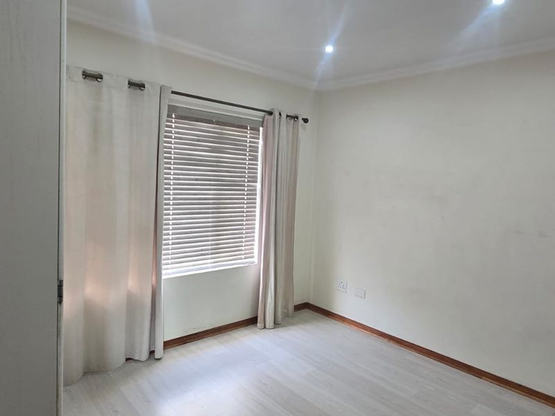 A bright, empty bedroom featuring light-toned laminate flooring, recessed ceiling lighting, and window treatments including venetian blinds and curtains.